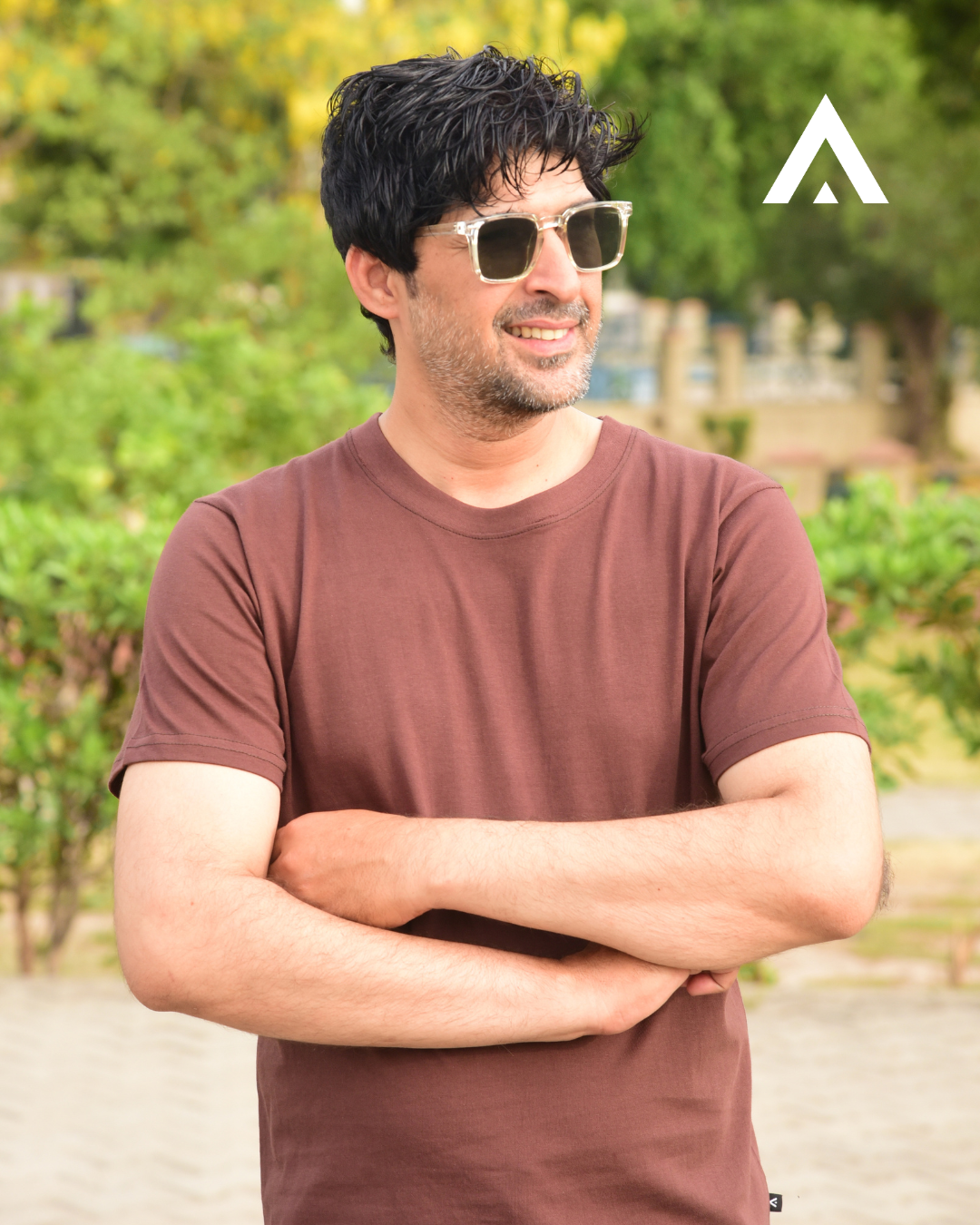 Man wearing sunglasses and a brown t-shirt with a blurred green outdoor background