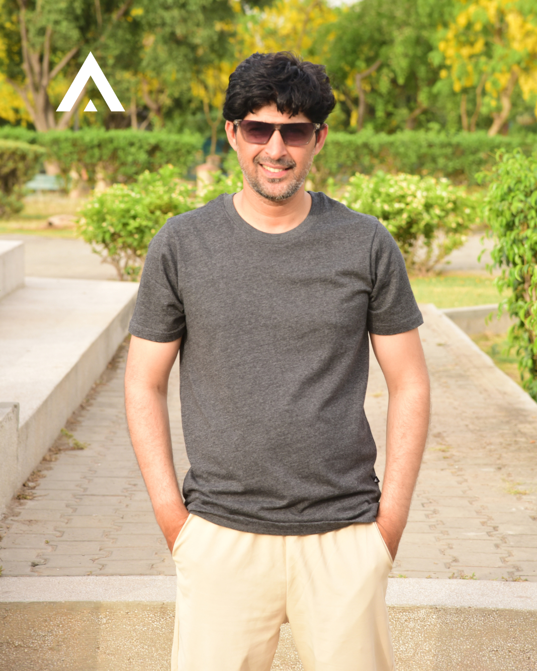 Man wearing sunglasses and a dark gray t-shirt in an outdoor setting with greenery.