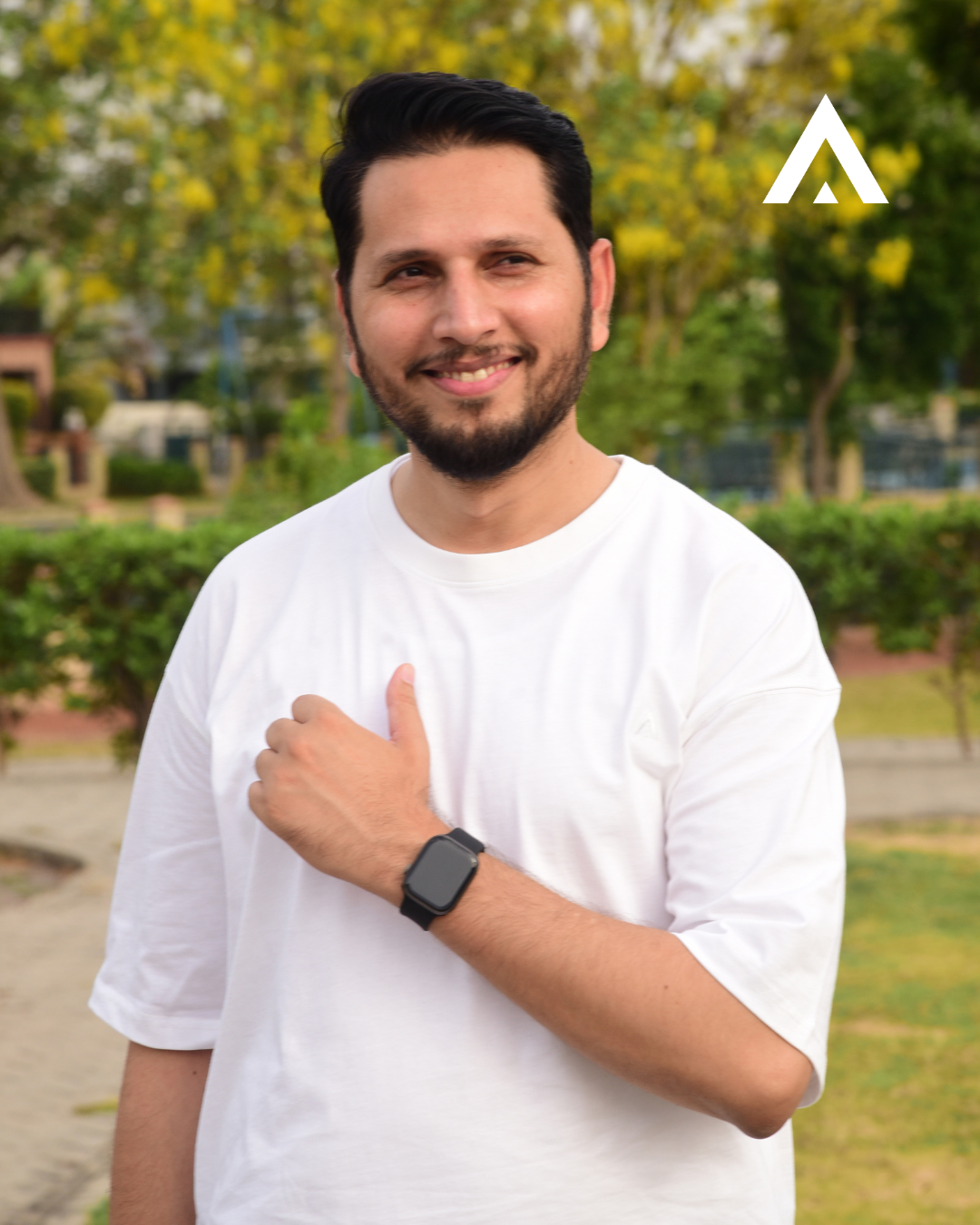 Man wearing a white t-shirt and black watch, standing outdoors with greenery in the background